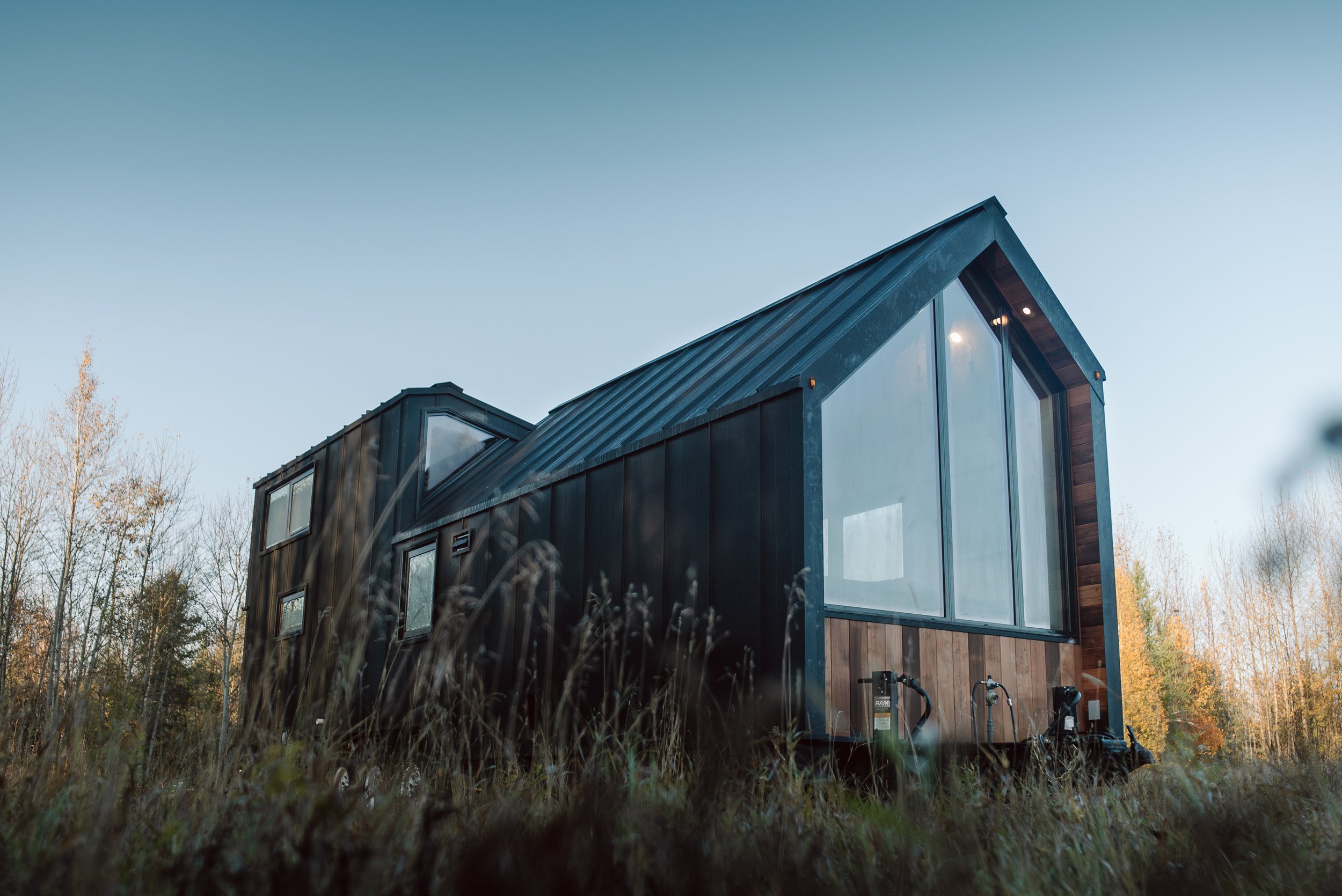 Modern house with large windows and black exterior, situated in a natural setting with trees in the background and grass in the foreground.